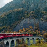 Red train traversing a scenic arch bridge in the Swiss Alps during autumn.